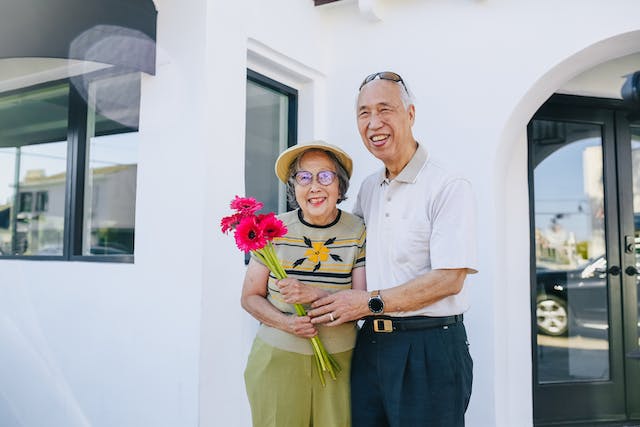 Happy old couple, holding a bouquet of bright red flowers and standing in front of a white home.